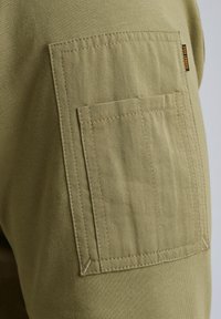 Il tessuto dei pantaloni cargo verde oliva presenta una superficie testurizzata, con una grande tasca laterale e dettagli di cuciture rinforzate.