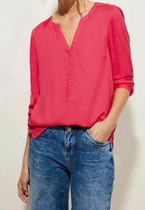 Persoon draagt een felroze blouse met lange mouwen en knopen en blauwe denim spijkerbroek, hand nonchalant in de zak.