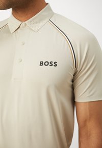 Béžová polo košela vyrobená z textúrovanej tkaniny, s golierom na gombíky, perforovanými detailmi a názvom značky "BOSS" v čiernej farbe.