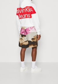 Sudadera blanca con un logo rojo y pantalones cortos negros que presentan un patrón texturizado en rosa, marrón y crema, combinados con zapatillas blancas.