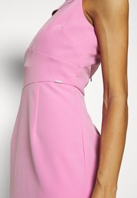 Robe rose sans manches présentant une coupe ajustée, des lignes épurées et un petit détail de label à la taille. Tissu lisse et léger.