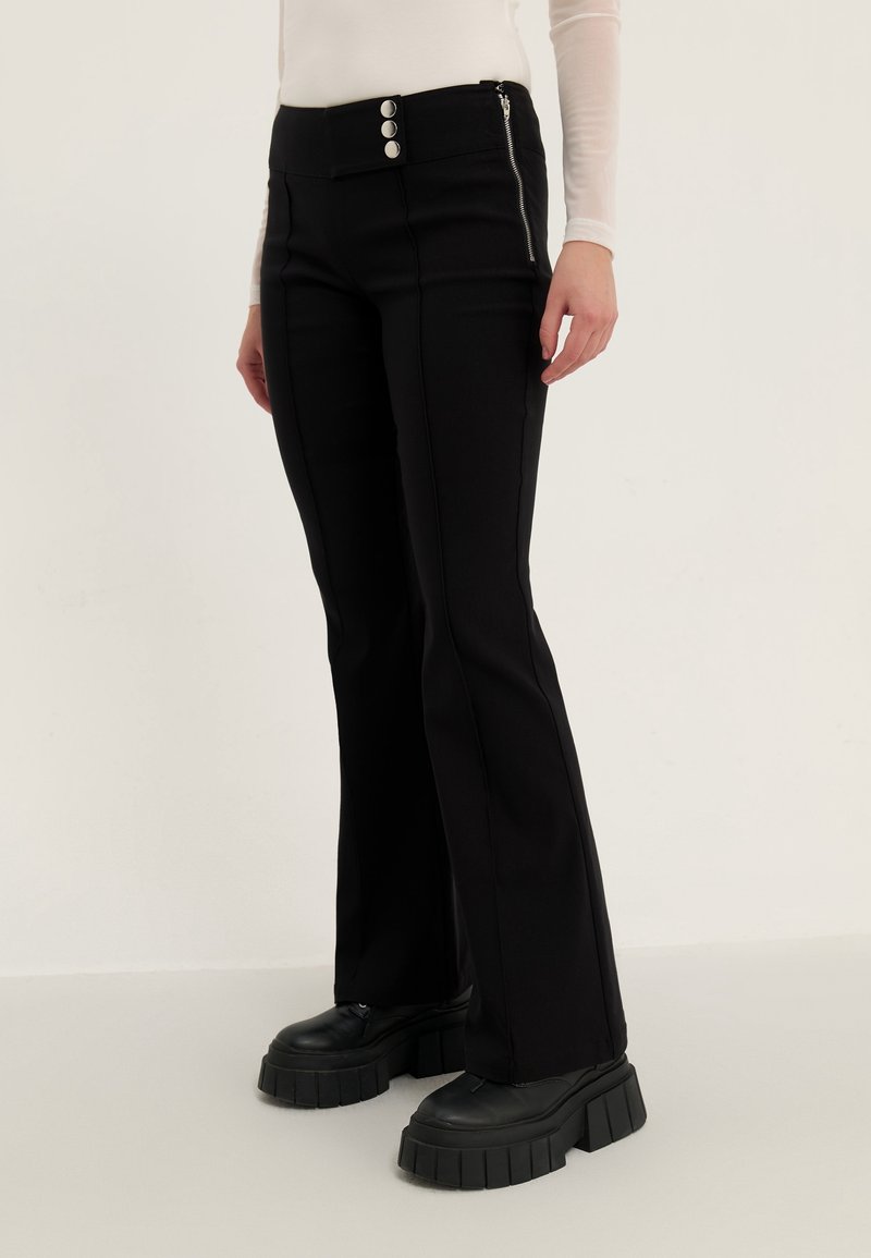 Personne portant un pantalon évasé noir avec détail à trois boutons et fermeture éclair latérale, associé à des chaussures plateformes noires grosses et un haut blanc à manches longues.