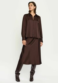 Rudos spalvos satininė palaidinė su sagomis priekyje ir siūta apykakle, derinama su to paties atspalvio ruda satininė sijonu. Modelis dėvi rudus kelio ilgio batus.