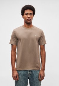 Άνδρας με κοντά σγουρά μαλλιά που φοράει μια ταμπά μπλούζα με μικρό λευκό λογότυπο στο στήθος και μπλε τζιν, στέκεται μπροστά σε απλό λευκό φόντο.