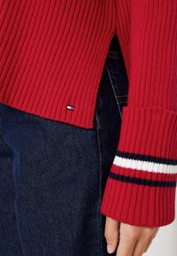 Czerwony sweter z żeberkowym wzorem, wykonany z teksturowanej tkaniny, z bocznym rozcięciem i paskowanymi mankietami w kolorze granatowym i białym. Na dolnym brzegu widoczna mała metka z logo.