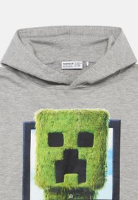 Γκρι φούτερ με κουκούλα που διαθέτει μια μεγάλη γραφική παράσταση πράσινου pixelated χαρακτήρα με μαύρα στοιχεία, περιτριγυρισμένη από μπλε φόντο. Το ύφασμα είναι μαλακό.