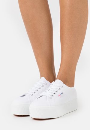 Białe trampki na platformie z zaokrąglonym czubkiem, wiązanym fasonem i teksturowaną gumową podeszwą. Detal z logo po zewnętrznej stronie.