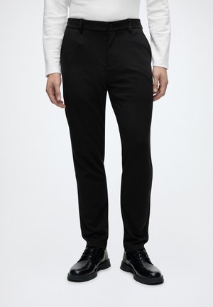 Mann trägt schwarze Slim-Fit-Hosen, schwarze Leder-Schnürschuhe und ein weißes langärmliges Oberteil, steht vor einem schlichten hellen Hintergrund.