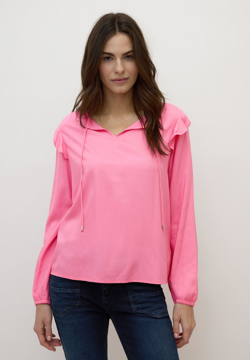 Blouse rose à manches longues avec une capuche, des épaules à volants et des cordons de serrage. Tissu lisse avec une coupe décontractée, portée sur un jean foncé.