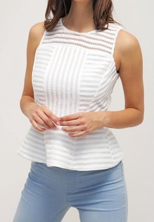 Hihaton valkoinen pitsipeplumtoppi, jossa pystysuora ja vaakasuora raidallinen kuvio, yhdistetty vaalean sinisten farkkujen kanssa, kädet ristissä vyötäröllä.