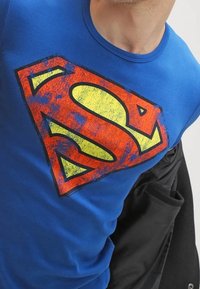 Niebieska bawełniana koszulka z dużym czerwono-żółtym logo Supermana o wyblakłej fakturze. Okrągły dekolt i krótkie rękawy.
