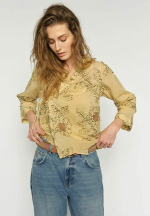 Nő egy sárga áttetsző virágos blúz szegélyét igazítja, kék farmernadrágot és barna övet visel, egyszínű háttér előtt.