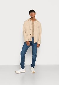 Modelo masculino em pé, vestindo uma jaqueta bege sobre uma camisa dourada, jeans azuis e tênis brancos, contra um fundo branco liso.