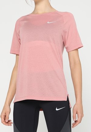 Kobieta w różowej sportowej koszulce Nike z krótkim rękawem i czarnych legginsach z logo Nike, stoi na prostym jasnym tle.