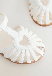 White leather sandals with horizontal straps and a heart cutout detail. Soft inner sole features floral pattern, complemented by a tan outsole.