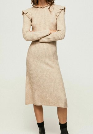 Frau trägt ein beiges, gestricktes Midikleid mit Zopfmuster, mit gerafften Schulterdetails und schwarzen Knöchelstiefeln, steht mit verschränkten Armen.