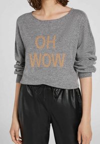 Donna che indossa un maglione grigio a maglia con "OH WOW" in lettere beige e pantaloni con cordoncino in finta pelle nera, braccia rilassate ai fianchi.