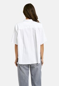 White short-sleeve shirt with a back pleat detail, made of cotton, featuring a button-down collar and a relaxed fit design.