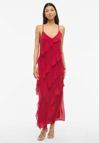 Raudona šifono maksi suknelė su voliotais sluoksniais, plonomis petnešėlėmis ir laisva siluetu. Sidabrinės šlepetės puikiai dera prie aprangos.