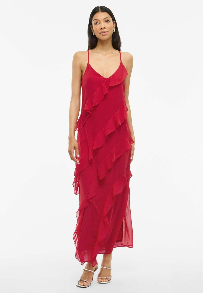 Raudona šifono maksi suknelė su voliotais sluoksniais, plonomis petnešėlėmis ir laisva siluetu. Sidabrinės šlepetės puikiai dera prie aprangos.