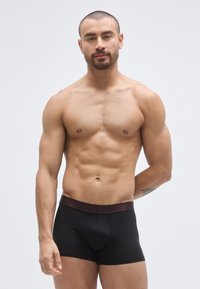 Černé boxerky s tmavě červeným páskem s logem, mají přiléhavý střih a elastický materiál pro pohodlí. Hladká textura, minimalistický styl.