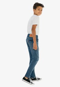 Jeans de mezclilla azul con un diseño de pierna recta, cintura alta y parche de marca de cuero. Combinados con una camiseta blanca básica y zapatillas negras.