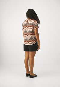 Short-sleeved top with a zigzag pattern in orange, white, and black; paired with black shorts and flat shoes. The back shows a rounded hem.