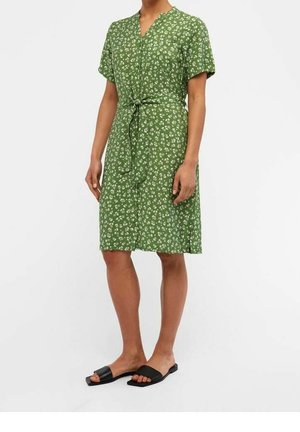 Grøn kjole med blomstermønster, korte ærmer, V-hals og taljebind; parret med sorte slidesandaler. Glat stofstruktur.