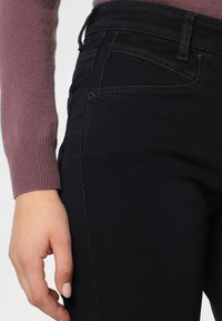 Ruka ženy odpočívající vedle černých džín a mauve svetr s dlouhým rukávem, se zaměřením na detaily boků a kapsy.