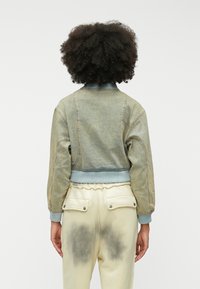 Zadnji pogled na kratko, svetlo zeleno bomber jakno z rebrastimi manšetami in ovratnikom, v kombinaciji s smetanastimi hlačami, ki imajo dva zadnja žepa.