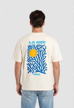 Hombre que lleva una camiseta crema con un diseño de coral azul y un círculo amarillo con el texto "LA MER" y "HERO7" en la espalda.
