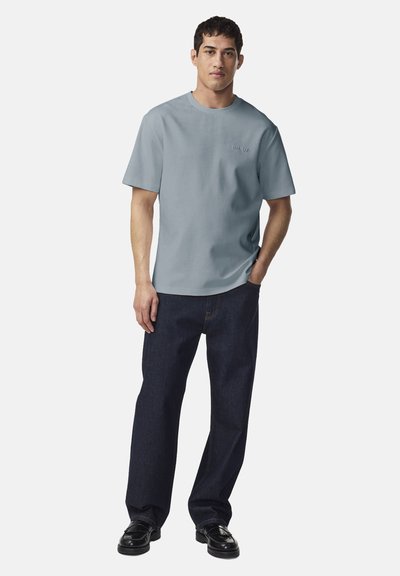 Mężczyzna ubrany w jasnoniebieską koszulkę z krótkim rękawem, ciemnoniebieskie dżinsy i czarne buty, stojący z jedną ręką w kieszeni na białym tle.