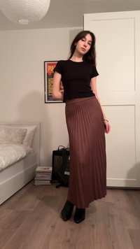 Jupe longue plissée marron associée à un T-shirt noir ajusté et des bottes à talons noires, posée sur un sol en bois clair avec un lit à la texture douce.