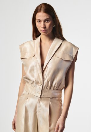 Femme aux longs cheveux roux portant un gilet beige en satin sans manches et un pantalon taille haute assorti, debout devant un fond uni.