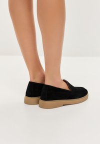 Czarne zamszowe buty wsuwane o niskim profilu z tanowym gumowym spodem, charakteryzujące się gładkim wykończeniem i minimalistycznym designem.
