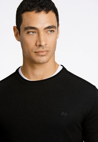 Maglione nero lavorato a maglia con scollo rotondo, caratterizzato da un sottile dettaglio logo sul petto e una texture a coste. Indossato sopra una maglietta bianca.