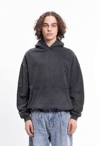 Hanorac supradimensionat de culoare gri închis, cu broderie pe buzunar și mâneci, realizat dintr-un material texturat și moale, cu un buzunar frontal tip „pouch”.