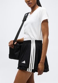 Γυναίκα που φοράει λευκό t-shirt και μαύρη αθλητική φούστα με λευκές ρίγες, κρατώντας μια μαύρη τσάντα χιαστί στον ώμο της.