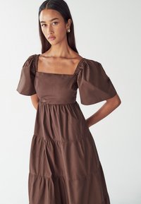 Ruda sluoksniuota suknelė su kvadratiniu iškirpimu, pūstomis trumpomis rankovėmis ir susiraukšlėjimais. Pagaminta iš lygaus, lengvo audinio su švelnia tekstūra.