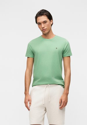 Junger Mann steht und trägt ein hellgrünes kurzärmeliges T-Shirt und cremefarbene Shorts mit Kordelzug vor einem schlichten weißen Hintergrund.