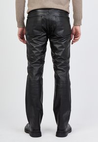 Schwarze Lederhosen mit glatter Textur, geradem Bein und zwei Gesäßtaschen. Das Modell trägt schwarze Schuhe, die den Saum zur Geltung bringen.