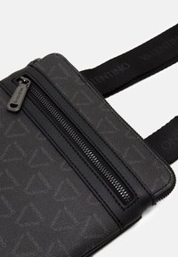 Black leather crossbody bag featuring a smooth finish, subtle logo pattern, front zip pocket, and adjustable webbing strap.
