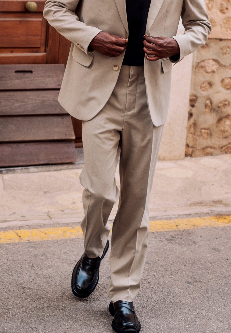 Mann in beige Sakko und Hose richtet beim Gehen auf der Straße sein Sakko, trägt schwarze Schuhe und ein dunkles Hemd darunter.
