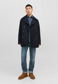 Giovane uomo in piedi che indossa un cappotto navy a doppio petto, camicia in denim blu, jeans blu e scarpe in camoscio marroni con lacci su sfondo grigio.
