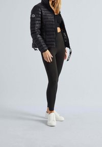 Sort smart jakke med lynlås og logo på ærmet, parret med sorte leggings og hvide sneakers, på en lys grå baggrund.