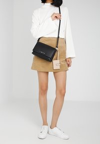 Žena v bílém tričku s dlouhým rukávem, světlé kordové mini sukni, bílých teniskách, držící černou crossbody kabelku s připojeným béžovým držákem na karty.