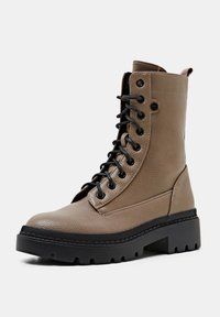 Botte de combat à lacets en cuir texturé taupe avec semelle noire épaisse à rainures et languette à l'arrière, inclinée pour montrer le côté extérieur.