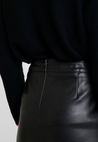 Detailní záběr osoby v černé kožené sukni a černém pleteném svetru se zipem na zadním pásku sukně.