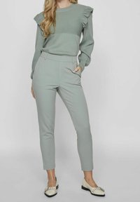 Vaaleanvihreä ribbitekoinen neule, jossa on rypytetty olkainyksityiskohta, yhdistettynä vaaleanvihreisiin räätälöityihin housuihin. Asun viimeistelevät musta-valkoiset balettitossut.