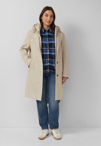 Beige gewatteerde jas met textuurstof, voorzakken en twee knopen. Paar blauwe jeans en witte sneakers met beige accenten.